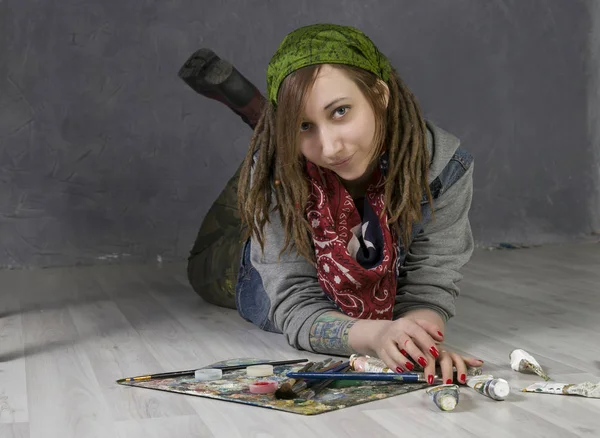 Atraktivní mladá zpěvačka s dredy leží na podlaze, drží štětce, jsou trubky s barvami v okolí — Stock fotografie