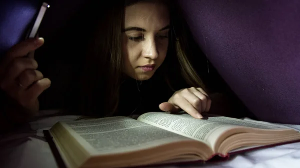Junge Frau versteckt sich unter einer Decke und liest nachts begeistert interessante Bücher. Mädchen zündet mit dem Handy als Taschenlampe — Stockfoto