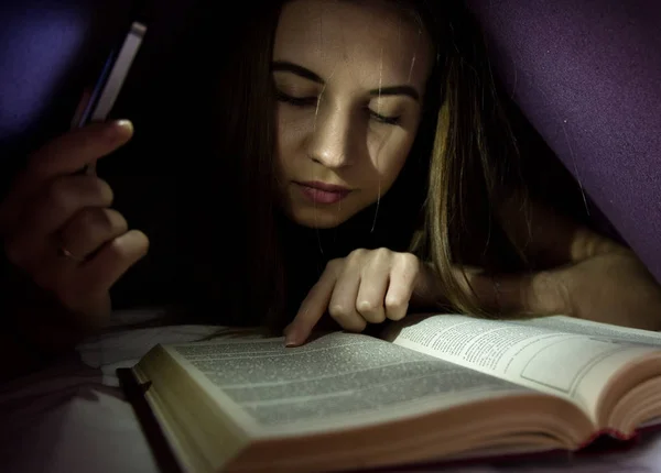 Genç kadın battaniye ve enrapt okuma kitabı geceleyin ilginç altında saklanıyor. Kız bir el feneri olarak telefon ile aydınlatma