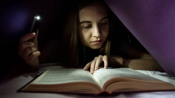 Νεαρή γυναίκα κρύβονται κάτω από την κουβέρτα και enrapt ανάγνωση ενδιαφέρον βιβλίο κατά τη διάρκεια της νύχτας. Κορίτσι φωτισμού με το τηλέφωνο ως φακό — Φωτογραφία Αρχείου