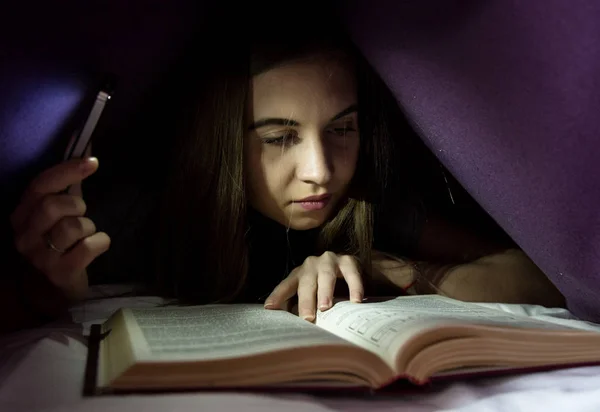 Junge Frau versteckt sich unter einer Decke und liest nachts begeistert interessante Bücher. Mädchen zündet mit dem Handy als Taschenlampe — Stockfoto