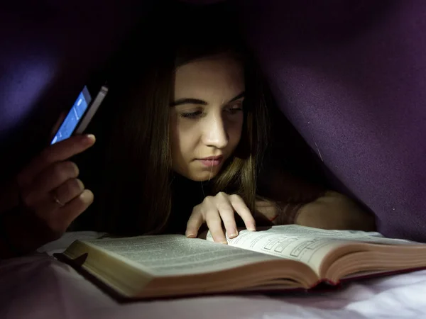 Νεαρή γυναίκα κρύβονται κάτω από την κουβέρτα και enrapt ανάγνωση ενδιαφέρον βιβλίο κατά τη διάρκεια της νύχτας. Κορίτσι φωτισμού με το τηλέφωνο ως φακό — Φωτογραφία Αρχείου