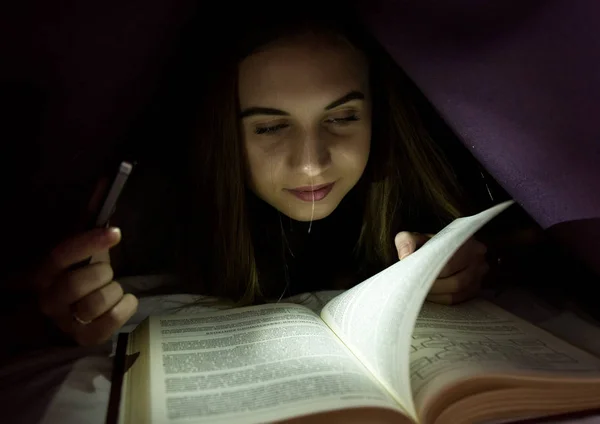 Mujer joven escondida debajo de una manta y enraptada leyendo un libro interesante por la noche. Chica de iluminación con el teléfono como una linterna — Foto de Stock