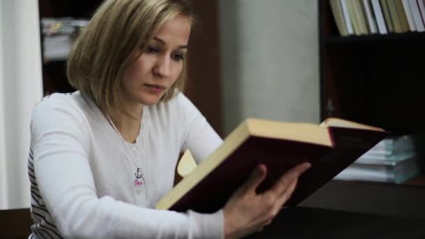 Una joven leyendo un gran libro en la biblioteca. estanterías con libros — Vídeo de stock