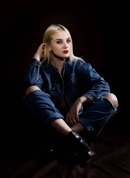 Divat stúdió fotó fiatal nő danim kabát és nadrág szakadt térdre ül a földön, sötét háttér. — Stock Fotó