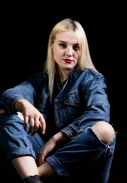 Mode-Studio-Foto einer jungen Frau in Danim-Jacke und -Hose mit zerrissenen Knien auf dem Boden sitzend, dunkler Hintergrund. — Stockfoto