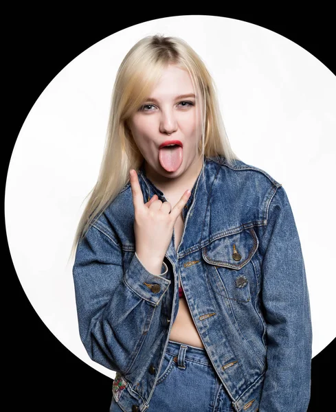 Young blonde woman in denim jaket and jeans shows tongue between two fingers on white background. indecent lifestyle concept. — Stock Photo, Image