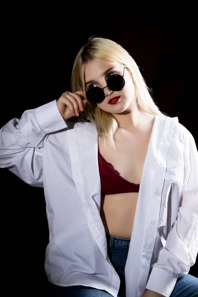 Pretty blond girl in a mans shirt and sunglasses posing on a dark background — Stock Photo, Image