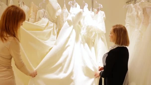 Jolie jeune femme choisissant une robe de mariée. assistant de magasin aide pour une fille dans le magasin de mariage . — Video