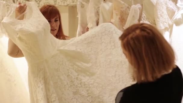 Помощник продавца помогает красивой молодой женщине выбрать свадебное платье в свадебном магазине — стоковое видео
