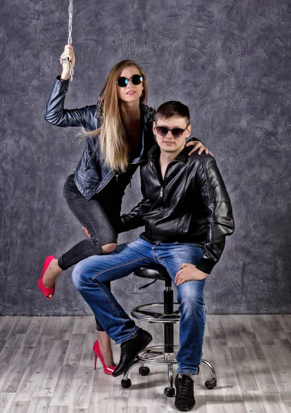 Young beautiful girl and handsome guy in leather jackets posing on a high chair on gray background — Stock Photo, Image