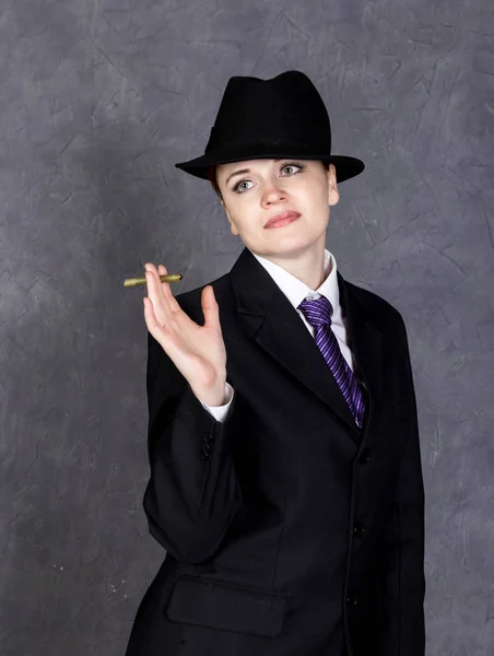 Mujer joven en estilo varonil con mini cigarro sobre fondo gris, chica en traje y corbata de hombre, camisa blanca y sombrero — Foto de Stock