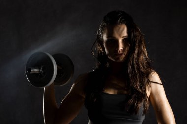 Koyu arkaplanda halterleri olan seksi bir kız. Sporcu spor salonunda egzersiz yapıyor.