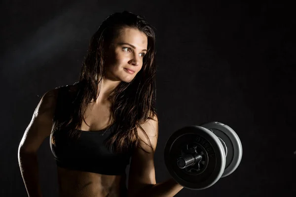 Fitness menina sexy com halteres em um fundo escuro. Atleta fazendo exercícios no ginásio — Fotografia de Stock