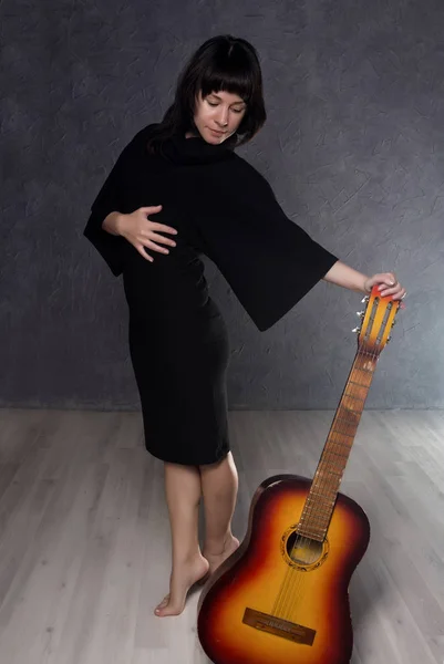Красива модна леді в готичній чорній сукні з високим коміром, позує з акустичною гітарою на сірому фоні . — стокове фото