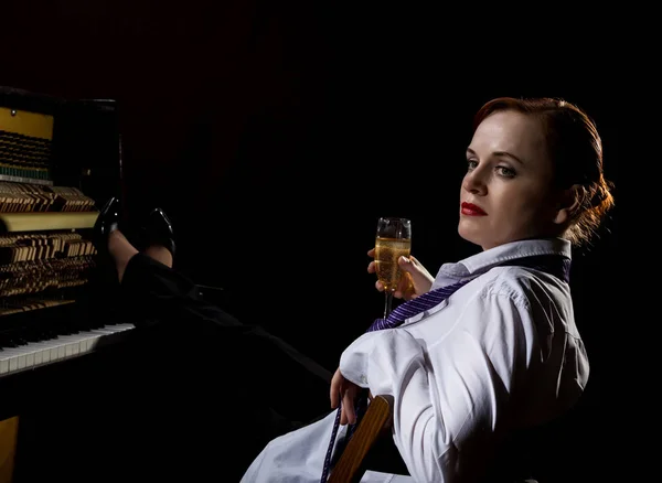 Элегантная женщина в белой рубашке и галстуке сидит рядом с пианино и пьет шампанское — стоковое фото