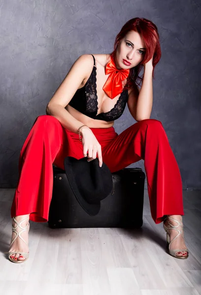 Woman in red pants and black hat with big boobs sits on a retro suitcase — Stock Photo, Image