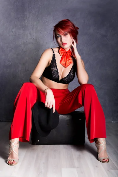 Woman in red pants and black hat with big boobs sits on a retro suitcase — Stock Photo, Image