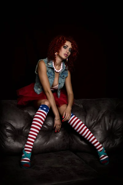 Curly redhead sexy girl sits on a leather sofa. Girl looks like a doll for adult men. Fashion style — Stock Photo, Image