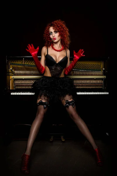 Ragazza sexy sembra una bambola con la rossa riccia in piedi accanto a un pianoforte, su uno sfondo scuro. Stile di moda — Foto Stock