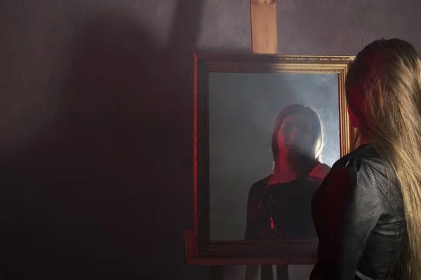 Hermosa mujer joven en una chaqueta de cuero se ve en el espejo como su retrato — Foto de Stock