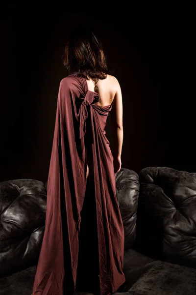 Glamour γυναίκα σε ένα καφέ cape stending σχετικά με ένα δερμάτινο καναπέ σε σκούρο φόντο. πίσω όψη — Φωτογραφία Αρχείου