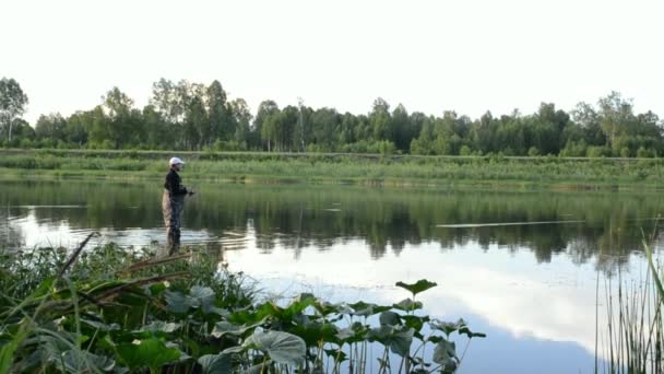 Рыбак рыбачит в спокойной реке утром. Человек в рыболовных снастях стоит в реке — стоковое видео