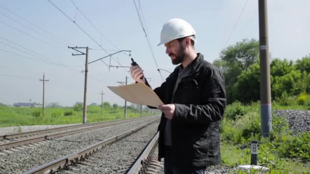 Inspektor ruchu kolejowego rozmawia walkie-talkie. Robotnik kolejowy w biały kask trzyma plan plany — Wideo stockowe