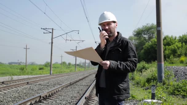 Inspector de tráfico ferroviario hablando irritablemente en walkie-talkie. Trabajador ferroviario en casco blanco con plano de planos — Vídeos de Stock