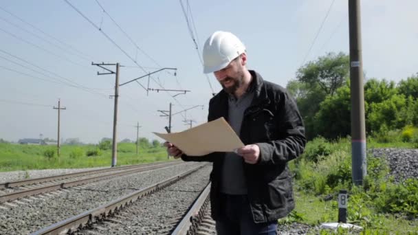 Inspetor de tráfego ferroviário irritantemente falando sobre walkie-talkie. Trabalhador ferroviário em capacete branco segurando plano de plantas — Vídeo de Stock