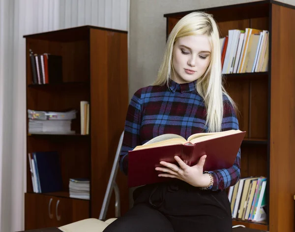 Νεαρό κορίτσι φοιτητής στέκεται και διαβάζοντας το βιβλίο κοντά σε ράφια στην βιβλιοθήκη φόντο — Φωτογραφία Αρχείου