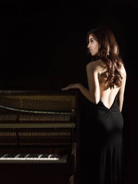 geri nere piyanonun karanlık bir arka plan üzerinde duran açık siyah elbiseli güzel genç kadın