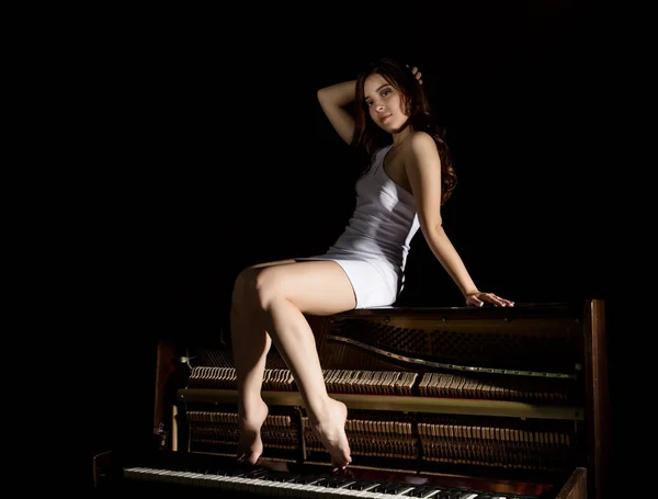 Hermosa mujer joven en un vestido blanco con una espalda abierta sentado en un piano viejo sobre un fondo oscuro — Foto de Stock