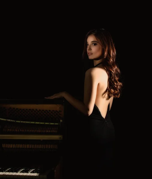 Όμορφη νεαρή γυναίκα σε ένα μαύρο φόρεμα με ανοιχτή πλάτη στέκεται nere πιάνο σε σκούρο φόντο — Φωτογραφία Αρχείου