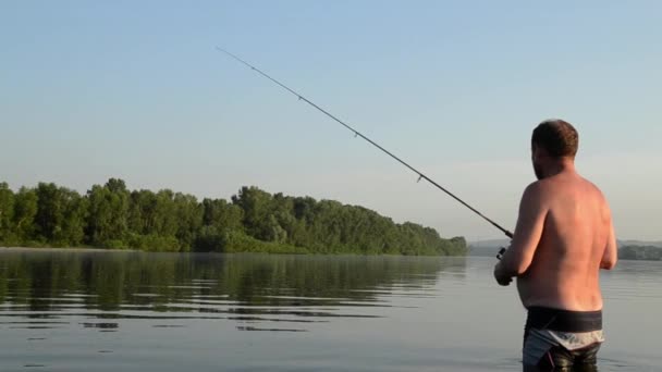 Рыбак рыбачит в спокойной реке утром. Человек в рыболовных снастях стоит в реке и бросает удочку — стоковое видео