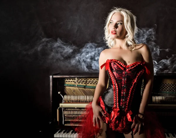 Gyönyörű szőke zenész piros fűző áll közel a zongora. Vintage stílusban gyönyörű nő. — Stock Fotó