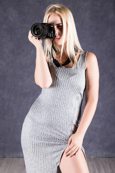 Mulher loira jovem em vestido cinza segurando câmera. Fotógrafo fazendo fotos . — Fotografia de Stock