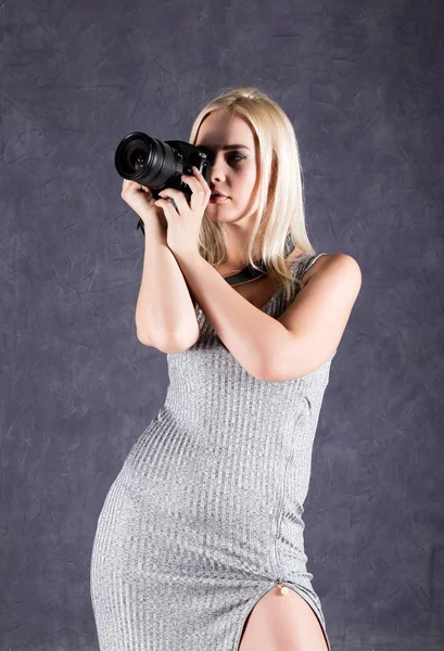 Молода блондинка в сірій сукні тримає камеру. Фотограф робить фотографії . — стокове фото