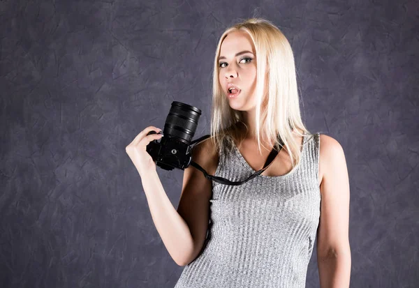 Mujer rubia joven en vestido gris sosteniendo la cámara. Fotógrafo haciendo fotos . — Foto de Stock