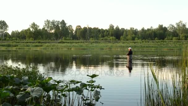 Рыбак рыбачит в спокойной реке утром. Человек в рыболовных снастях стоит в реке и бросает удочку — стоковое видео