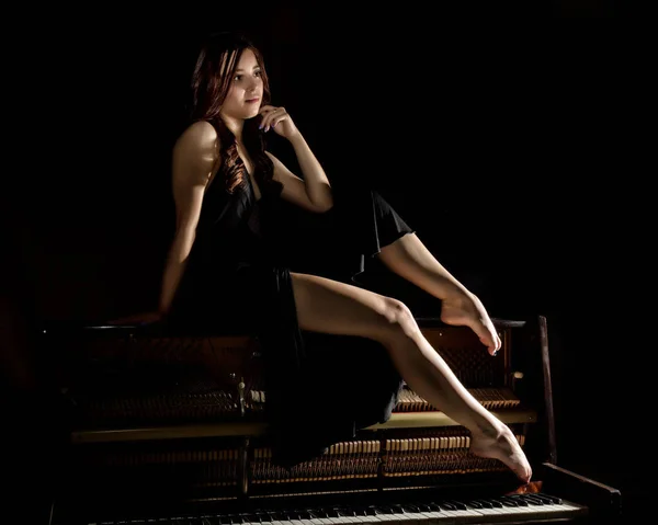 Beautiful young woman in a black dress with an open back sitting on a old piano on a dark background — Stock Photo, Image