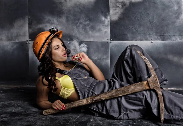 Beautiful sexy miner worker with a pickaxe and helmet sitting on a floor on steel background and smokes
