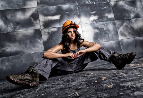 Красивий втомлений сексуальний робітник шахтаря в помаранчевому шоломі з ліхтариком, що сидить на підлозі на сталевому фоні — стокове фото