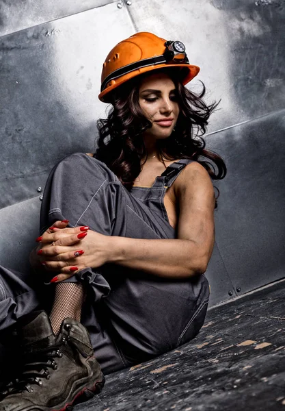 Beautiful tired sexy miner worker in orange helmet with flashlight sitting on a floor on steel background