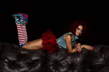 Kıvırcık saçlı seksi kızı deri kanepede oturuyor. Kız Yetişkin erkekler için bir bebek gibi görünüyor. Moda stil
