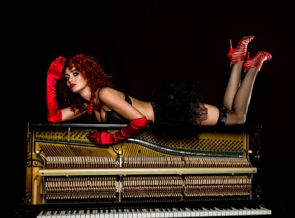Menina sexy se parece com uma boneca com ruiva encaracolado encontra-se em um piano, em um fundo preto. estilo de moda antiga — Fotografia de Stock