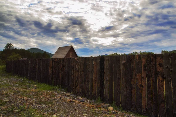 Pequeña casa junto a la antigua cerca de madera en el pueblo sobre un fondo de nubes oscuras — Foto de Stock