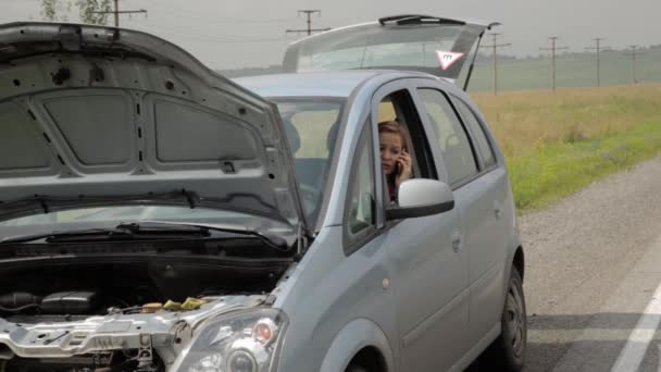 Ξανθιά κοπέλα κλήση κινητό τηλέφωνο σε έναν πανικό που κάθεται στο αυτοκίνητό της σπασμένα με ανοικτή κουκούλα σε έναν επαρχιακό δρόμο — Αρχείο Βίντεο