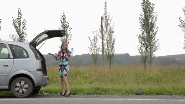 Chica rubia llamando al teléfono celular en un pánico cerca de su coche roto con tronco abierto en una carretera de campo — Vídeo de stock