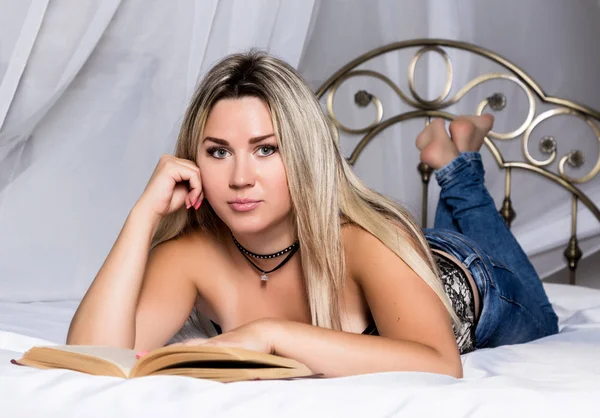 Mujer rubia acostada en la cama en una habitación luminosa y leyendo un libro —  Fotos de Stock